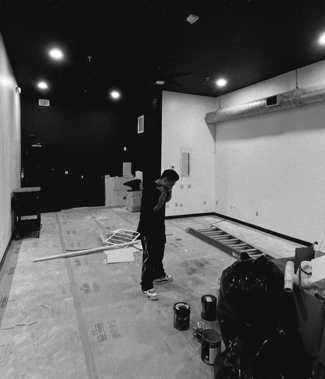 Javier alone in the shop under construction — black ceiling, white walls, paint cans on the floor.